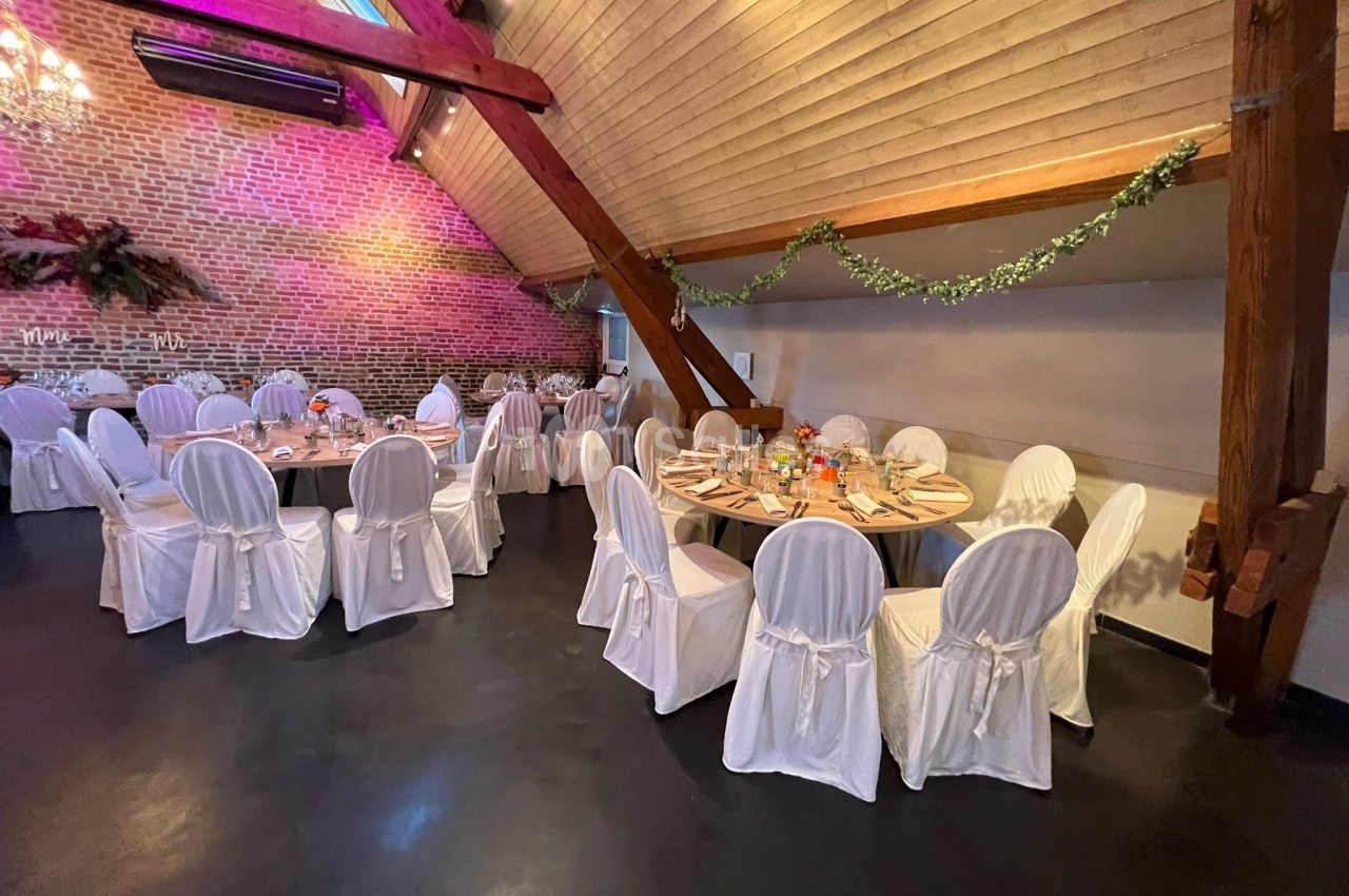 Salle de réception avec tables rondes dressées, chaises blanches et décoration florale sous un plafond en bois.
