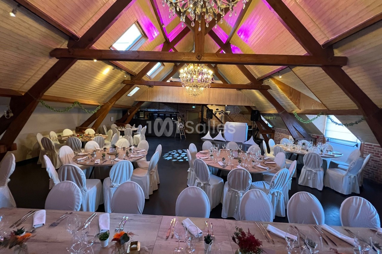 Salle de réception décorée avec des tables rondes dressées, des chaises blanches et un plafond en poutres apparentes.