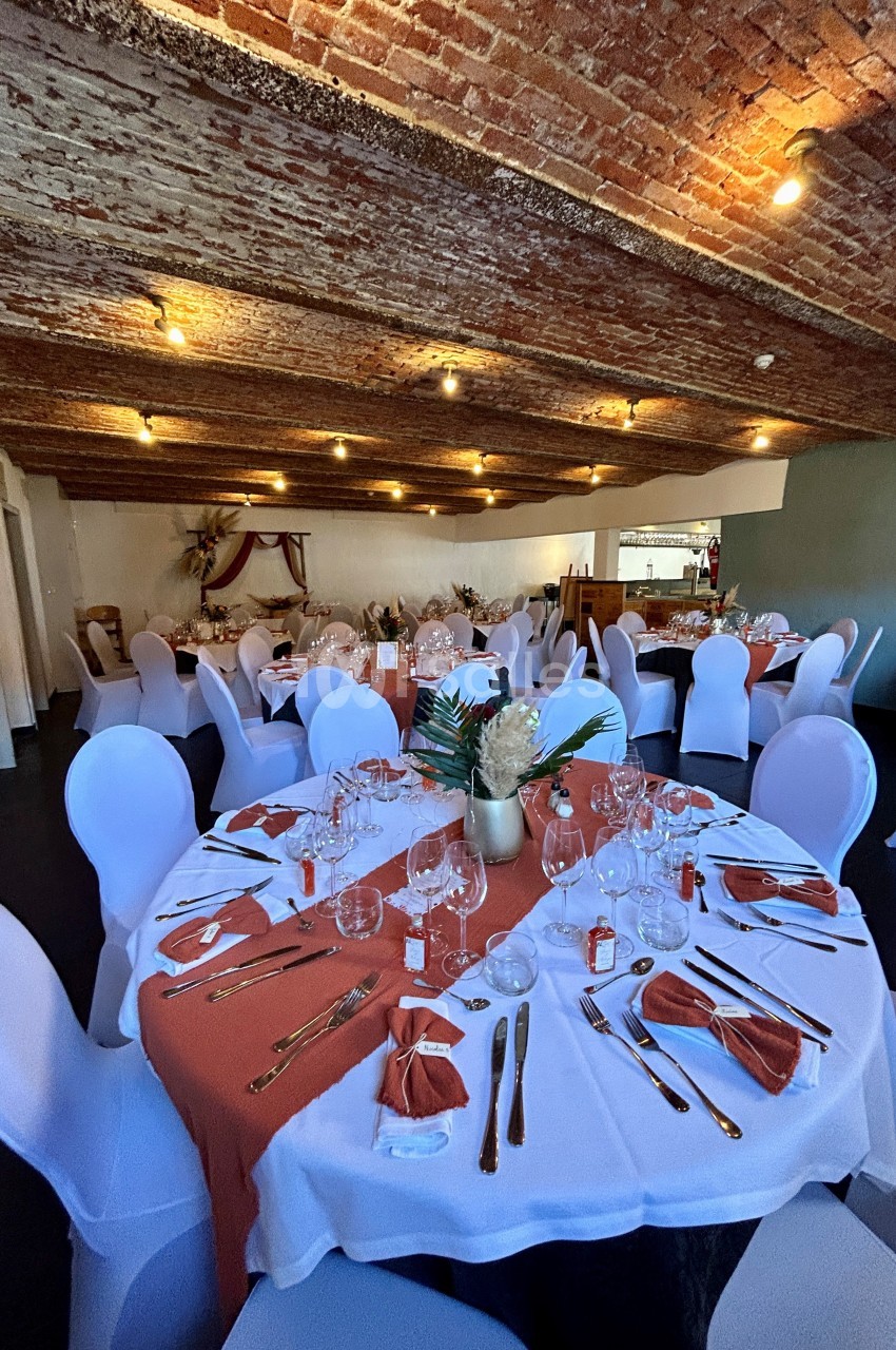 Salle de réception avec tables rondes dressées, nappes blanches, chemins de table orange et plafond en briques apparentes.