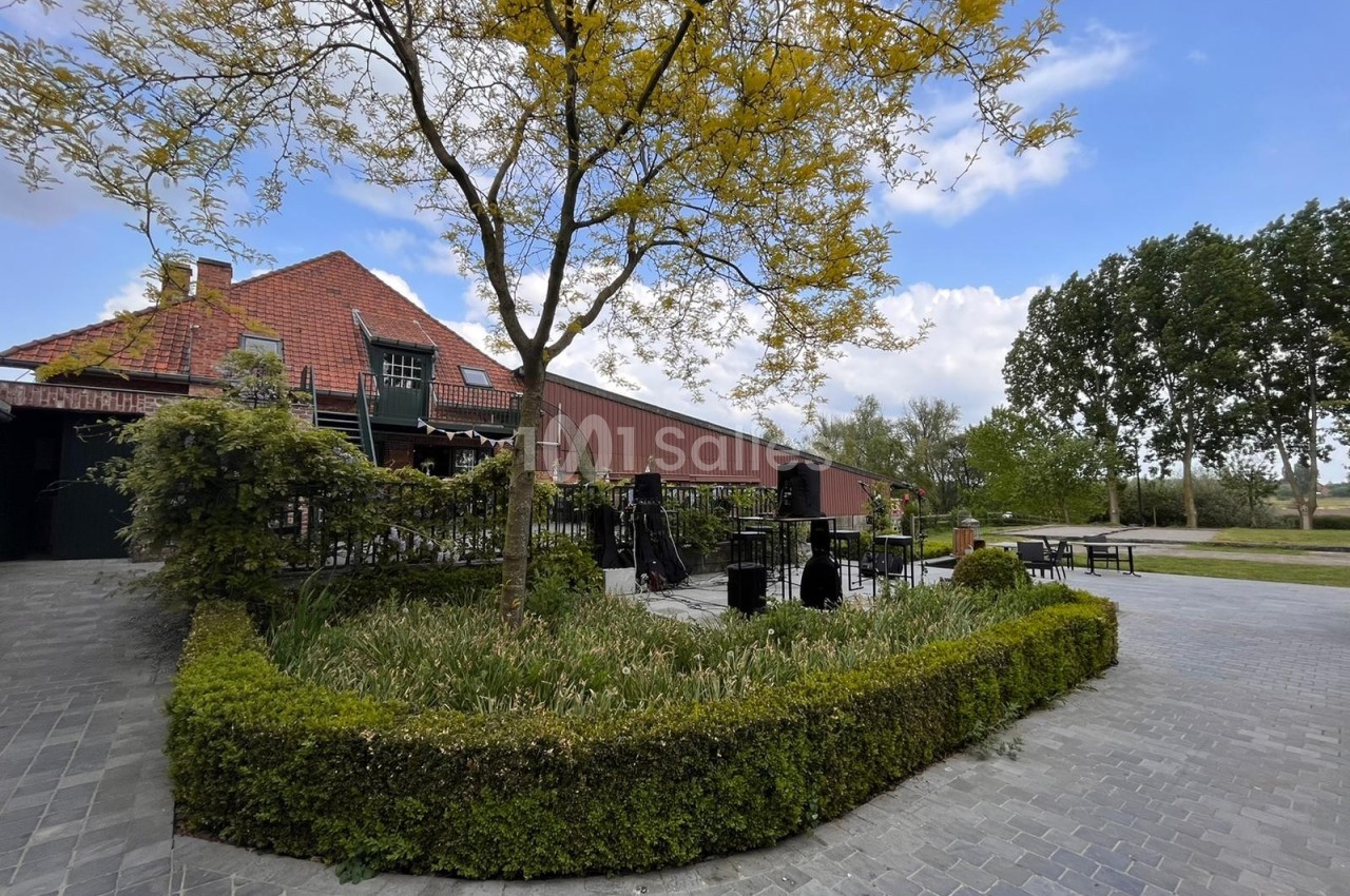 Terrasse extérieure avec mobilier sous un arbre, bordée de haies, près d'un bâtiment en briques rouges et d'un ciel dégagé.