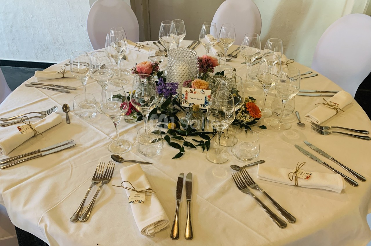 Table ronde élégamment dressée avec nappes blanches, couverts, verres, serviettes pliées et centre de table floral.