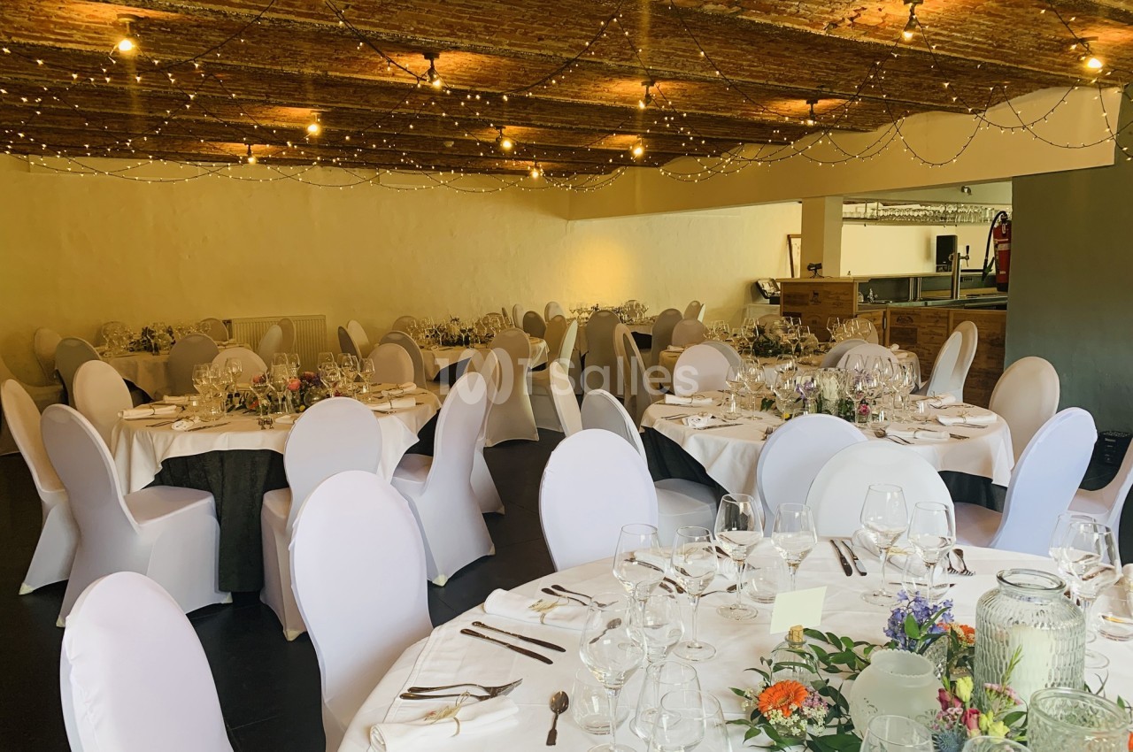 Salle de réception décorée avec des tables rondes dressées, nappes blanches, chaises houssées et guirlandes lumineuses au…