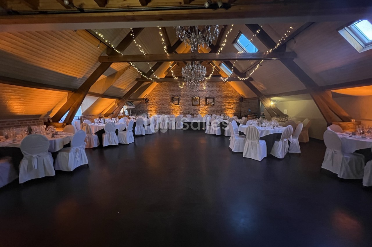 Salle de réception sous combles avec tables rondes dressées, chaises blanches et éclairage chaleureux.