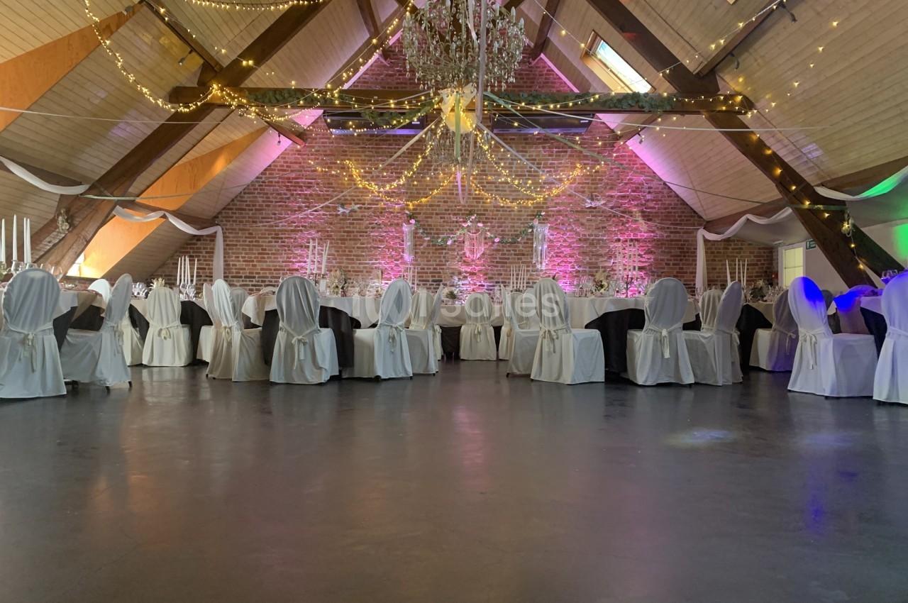 Salle de réception décorée avec des chaises habillées, tables dressées et guirlandes lumineuses suspendues.