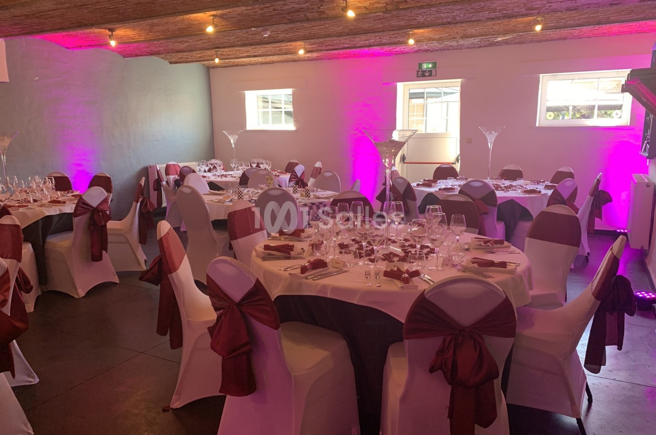 Salle de réception décorée avec des tables rondes dressées, nappes blanches, chaises ornées de nœuds rouges et éclairage…