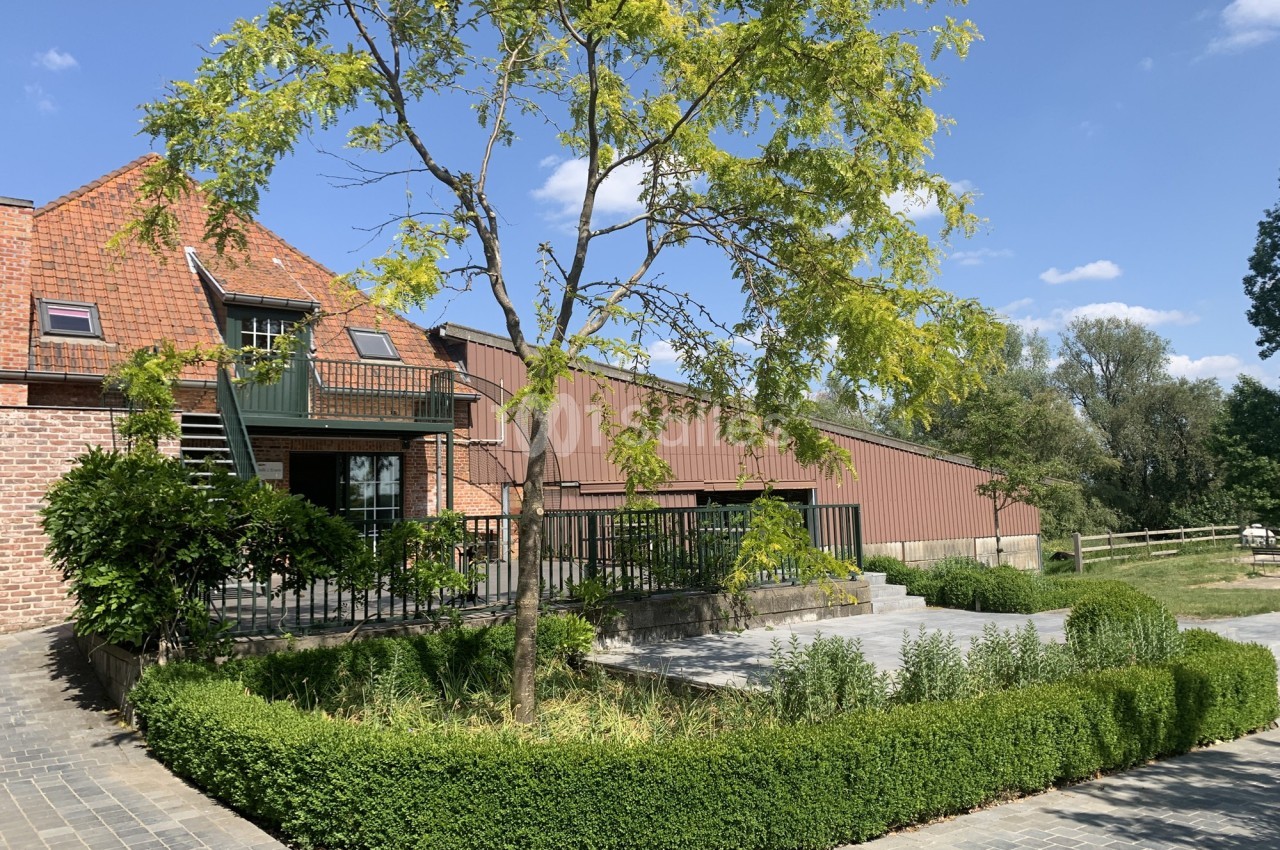 Bâtiment en briques et métal avec terrasse, entouré de verdure et d'un chemin pavé sous un ciel bleu.