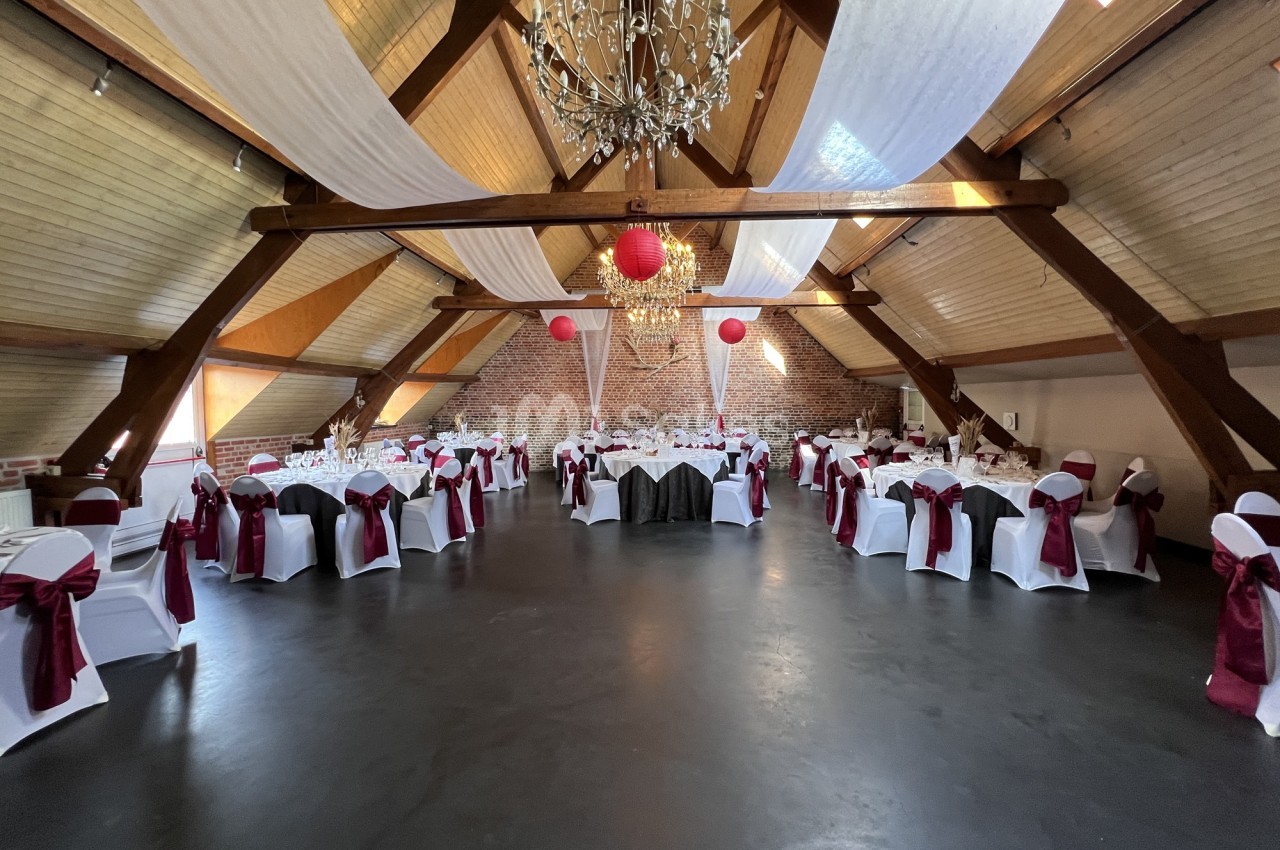 Salle de réception décorée avec des tables rondes, nappes blanches, chaises ornées de nœuds bordeaux et lustres suspendus.