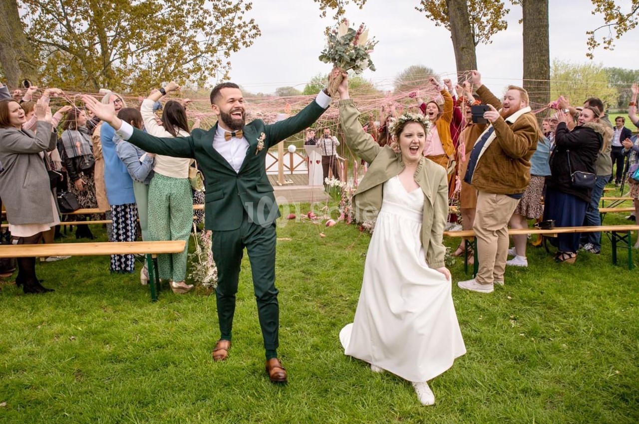 Un couple souriant en tenue de mariage marche sur une pelouse, entouré d'invités célébrant avec enthousiasme.