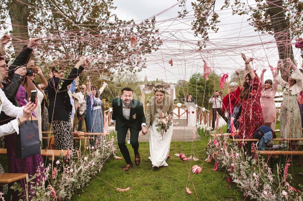 Un couple de mariés souriants traverse une allée décorée de fils colorés, entouré d'invités célébrant.