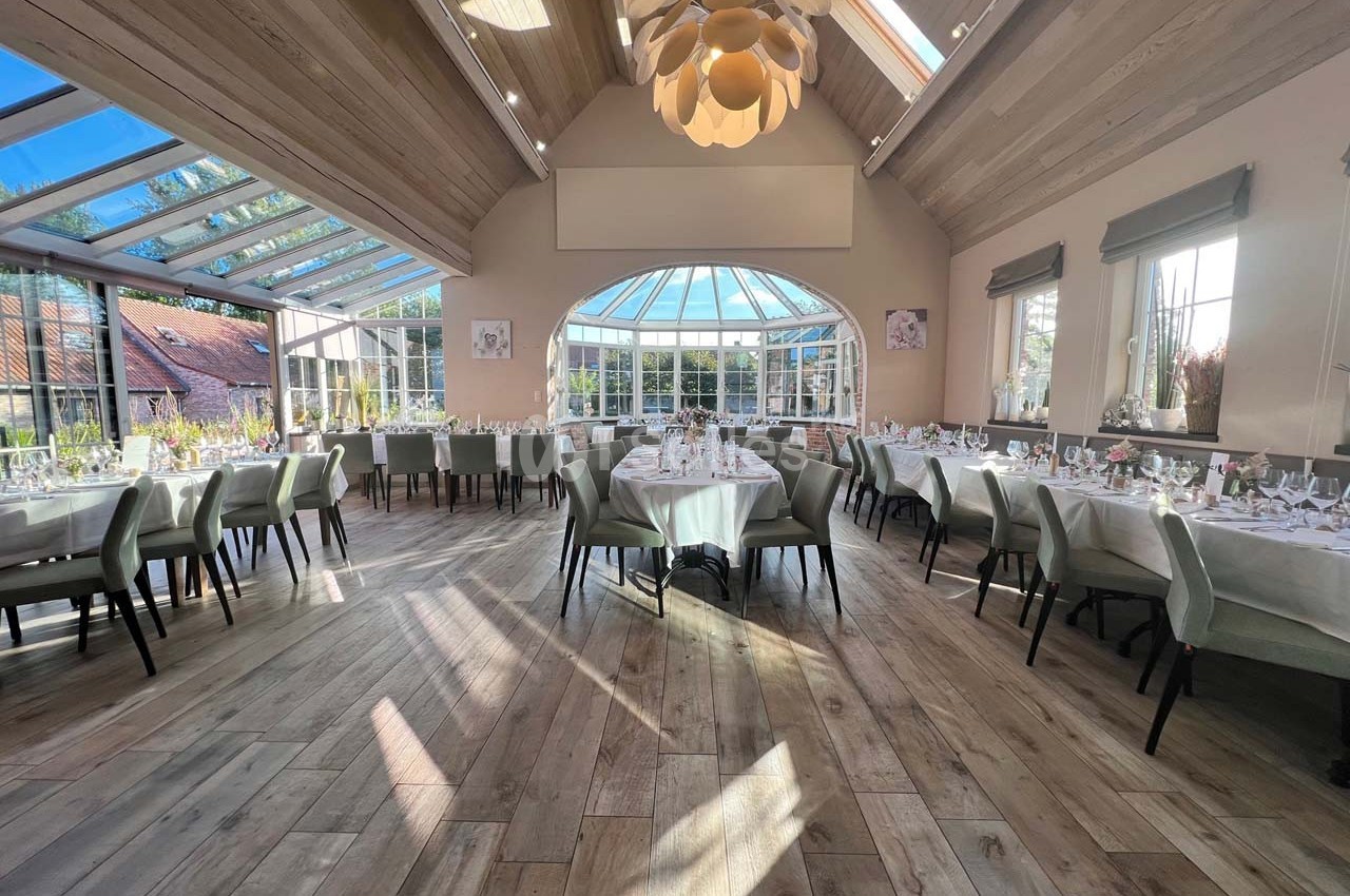 Salle lumineuse avec de grandes baies vitrées, tables dressées pour un repas, parquet en bois clair et plafond voûté.