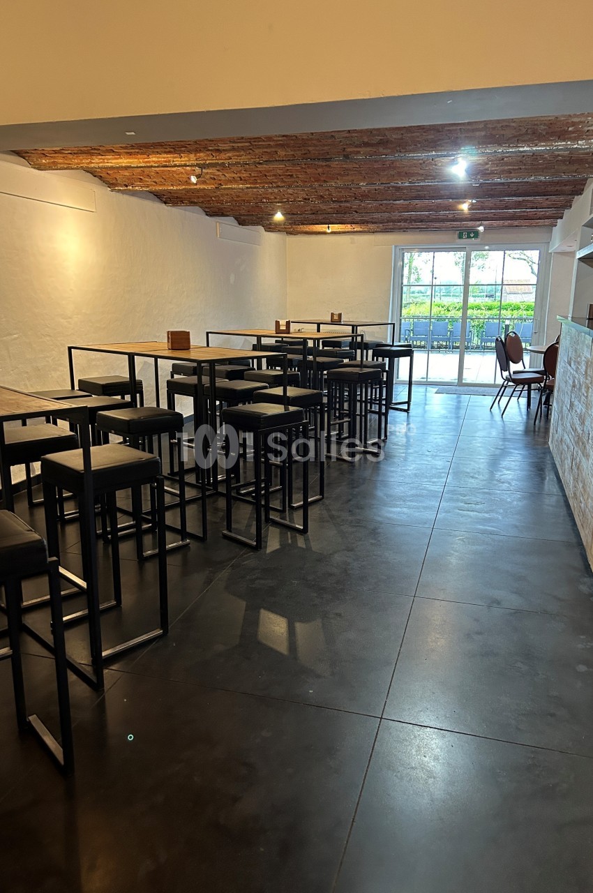 Salle lumineuse avec tables hautes, tabourets noirs, sol en béton et plafond en bois, donnant sur une baie vitrée.