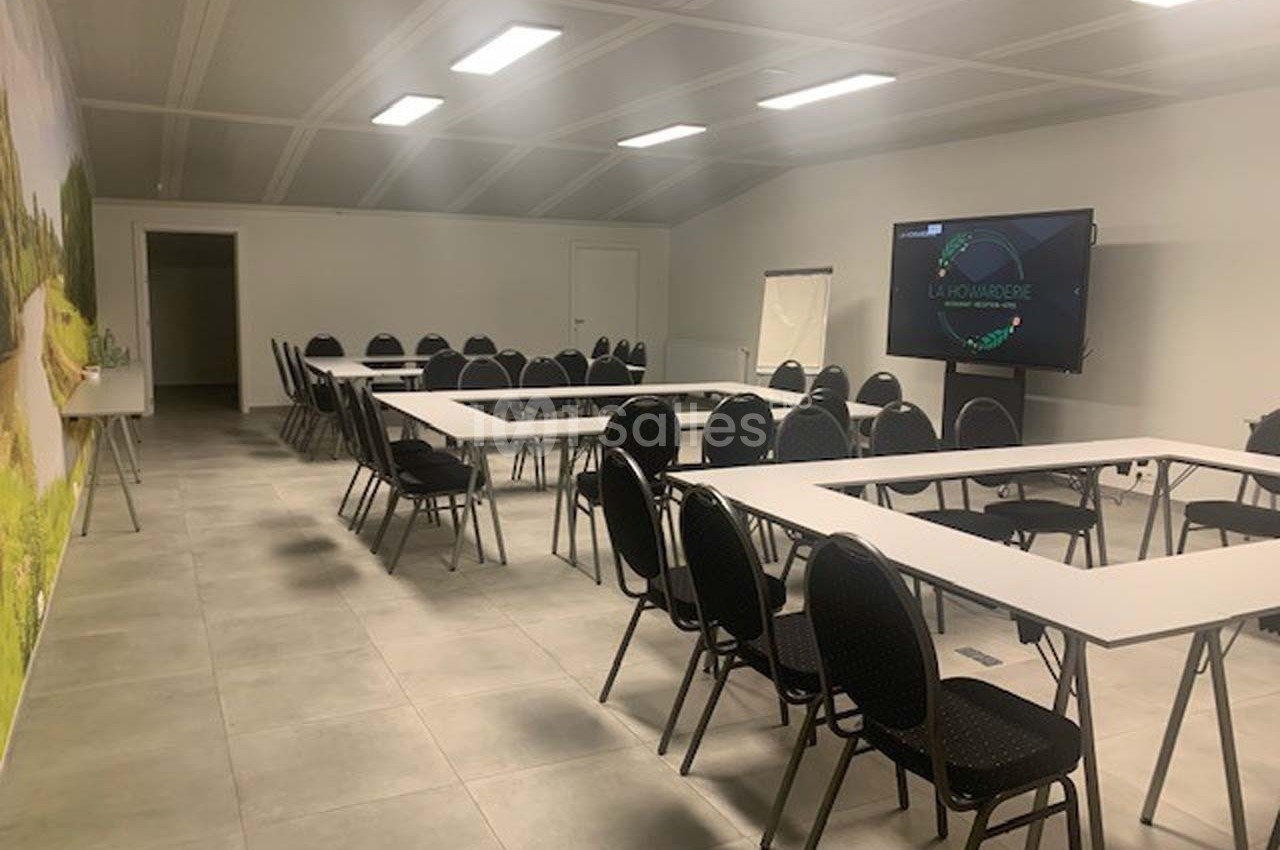 Salle de réunion lumineuse avec tables disposées en U, chaises noires, écran de présentation et tableau blanc.