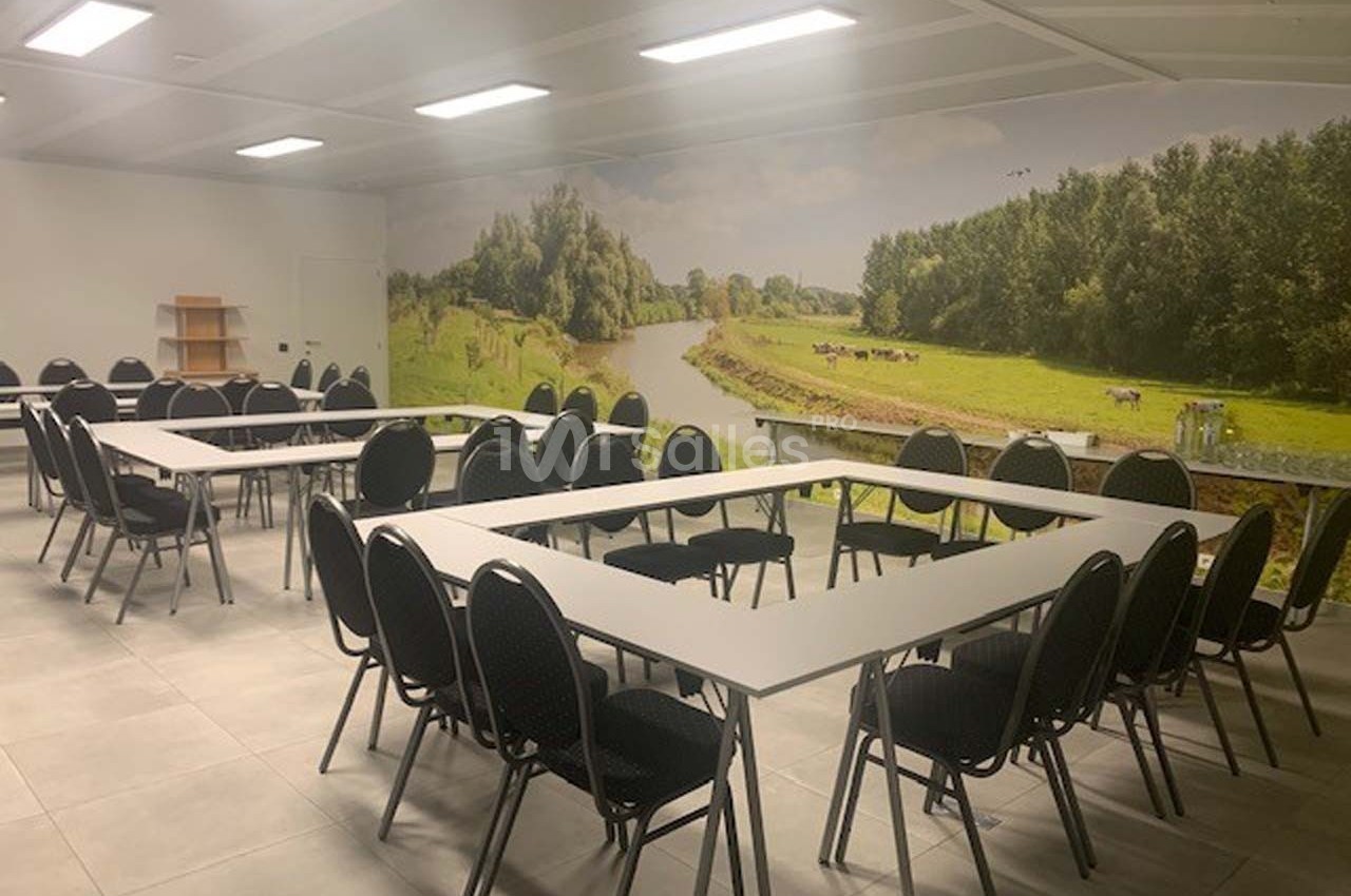 Salle de réunion lumineuse avec des tables disposées en U, des chaises noires et un mur décoré d'un paysage champêtre.