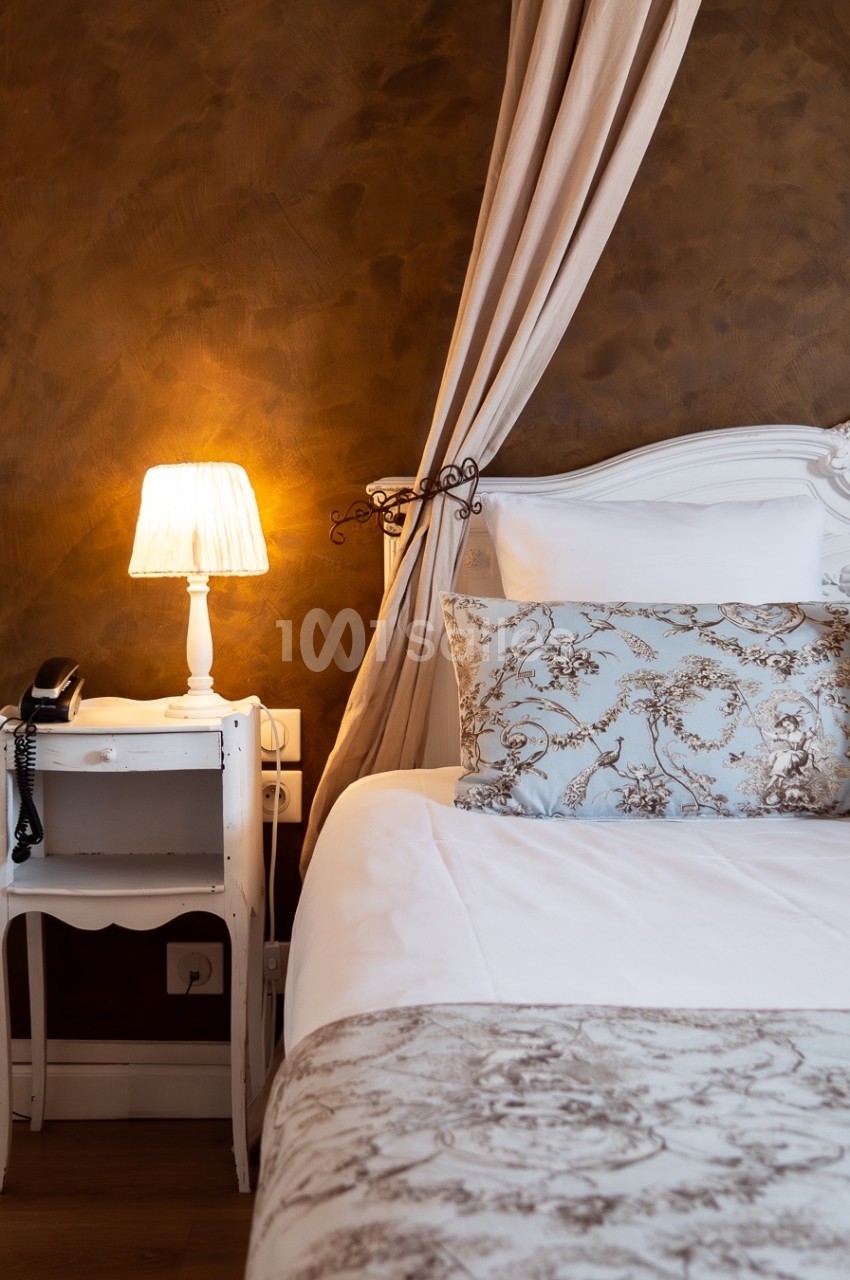 Lit avec tête de lit blanche, draps à motifs floraux, table de chevet avec lampe allumée et téléphone noir.
