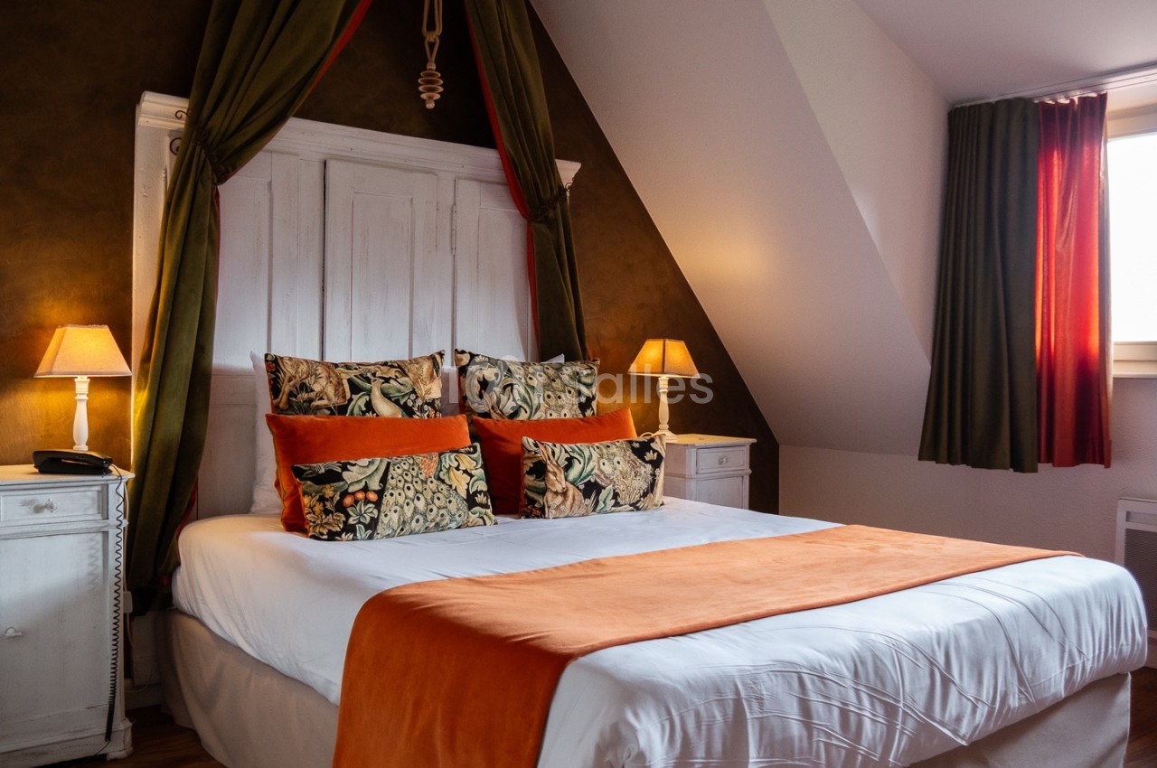 Lit double avec draps blancs, jeté de lit orange, coussins décoratifs et deux lampes de chevet dans une chambre chaleureuse.