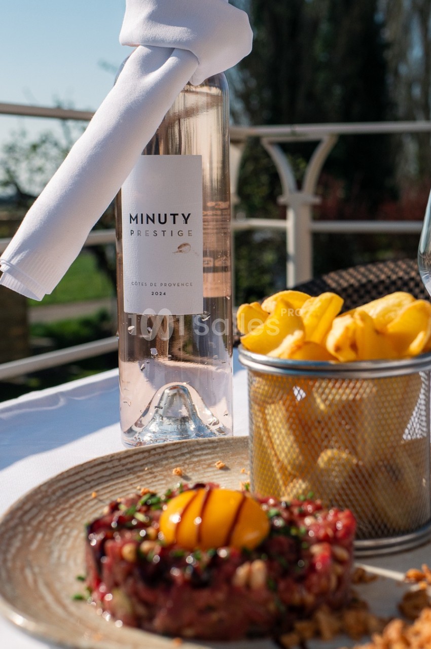 Bouteille de vin rosé, tartare de viande avec œuf, frites et verre sur une table en extérieur ensoleillé.