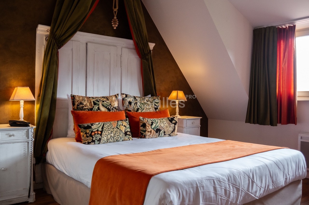 Lit double avec draps blancs, couverture orange, coussins décoratifs, encadré par deux lampes sur des tables de chevet…
