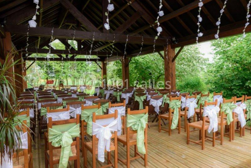 Chaises en bois décorées de rubans blancs et verts alignées sous une pergola en bois, entourée de verdure.