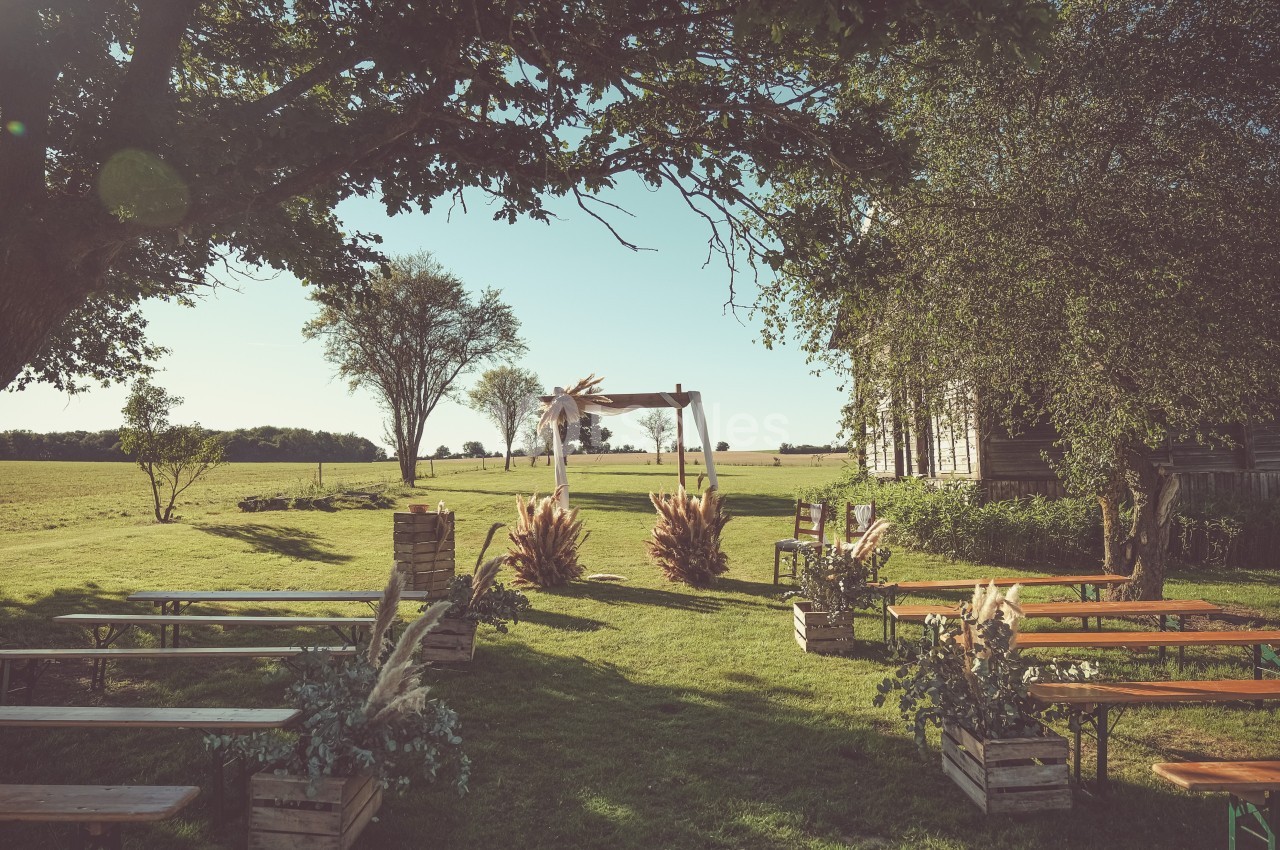 Cérémonie en plein air avec arche décorée de fleurs séchées, bancs en bois et paysage champêtre en arrière-plan.