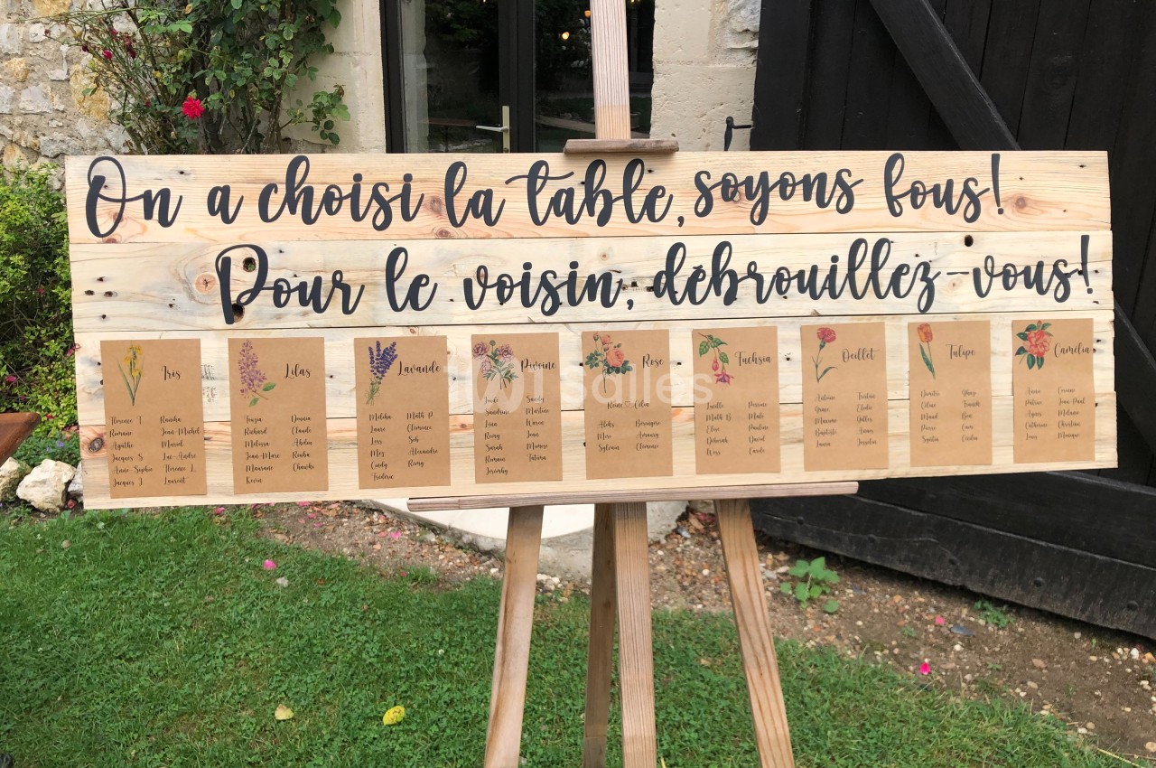 Panneau en bois avec des cartes de placement de table pour un événement, décoré de fleurs et d'une inscription humoristique.
