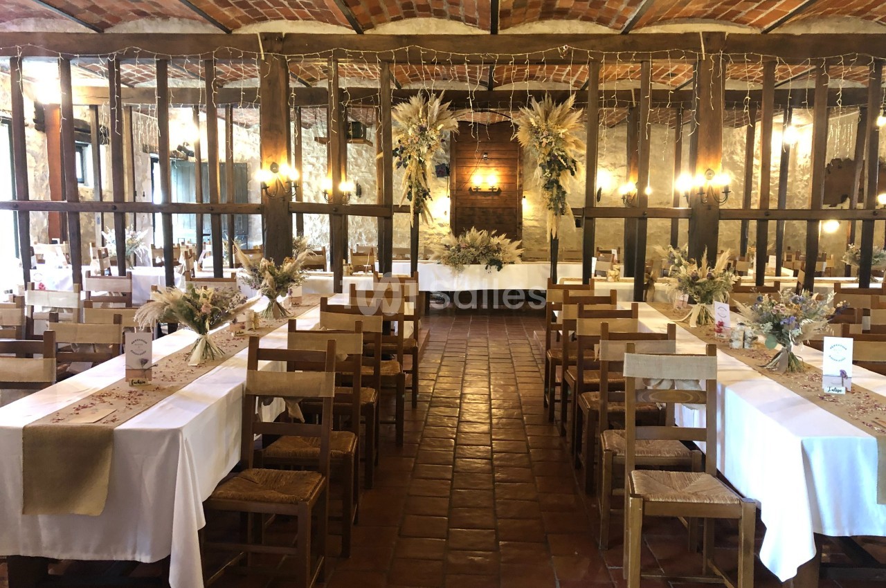 Salle de réception rustique avec tables décorées de nappes blanches, fleurs séchées et chaises en bois.