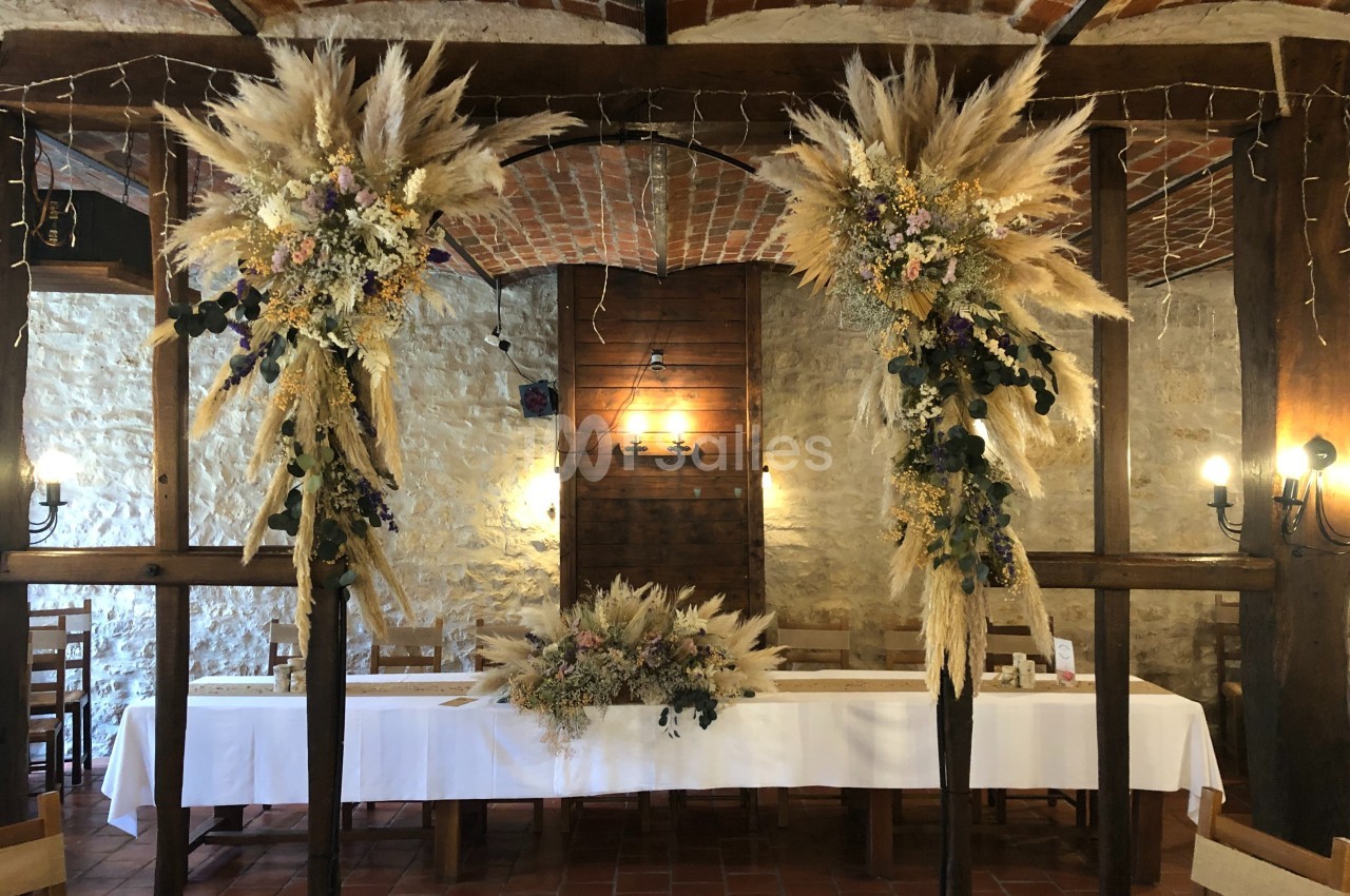 Décoration florale avec pampas et fleurs séchées encadrant une table blanche dans une salle en pierre et bois.