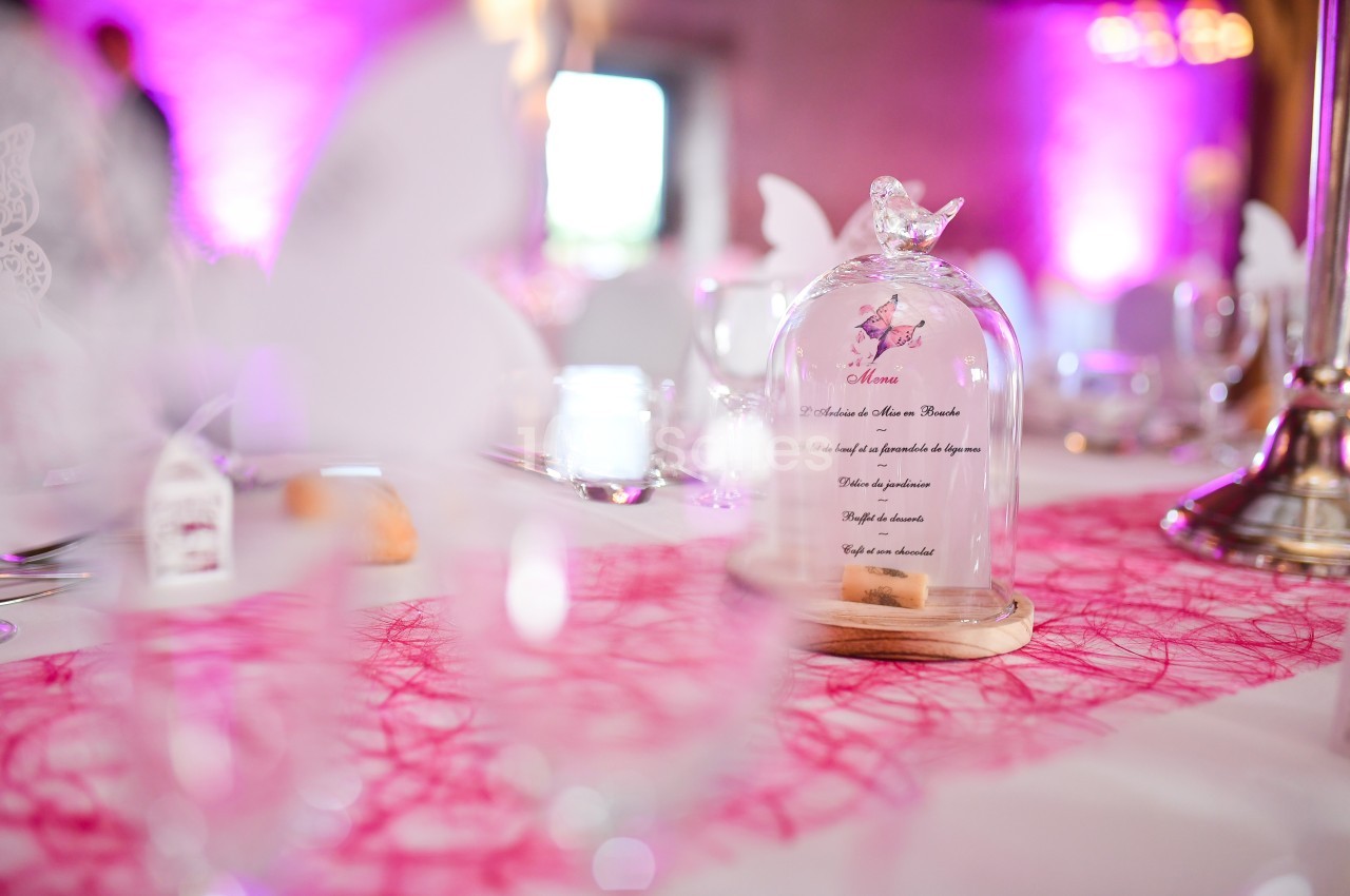 Cloche en verre contenant un menu décoratif sur une table de réception avec nappe rose et lumière tamisée.