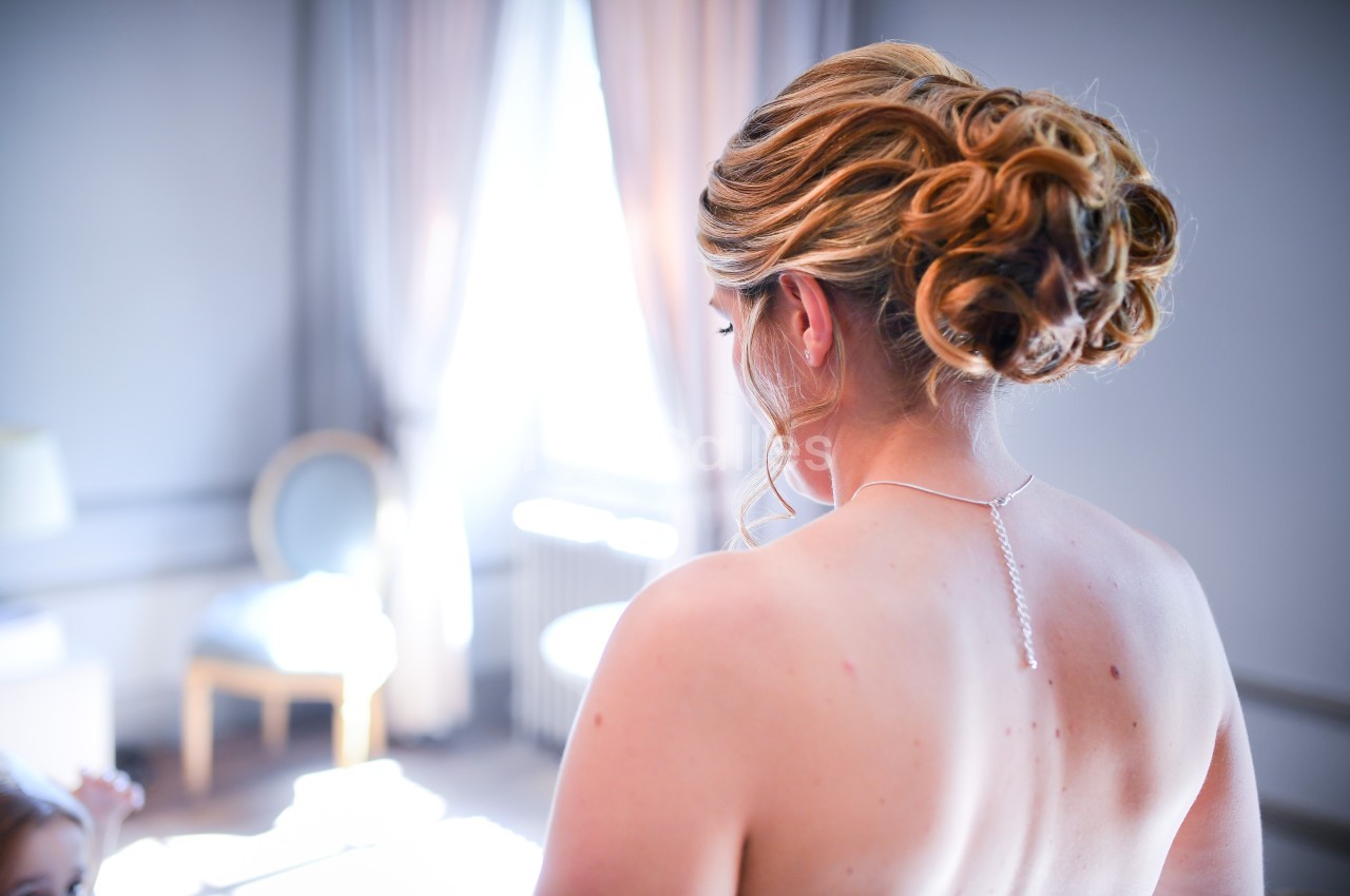 Femme de dos avec un chignon élaboré, portant un collier fin, dans une pièce lumineuse avec des rideaux et un fauteuil.