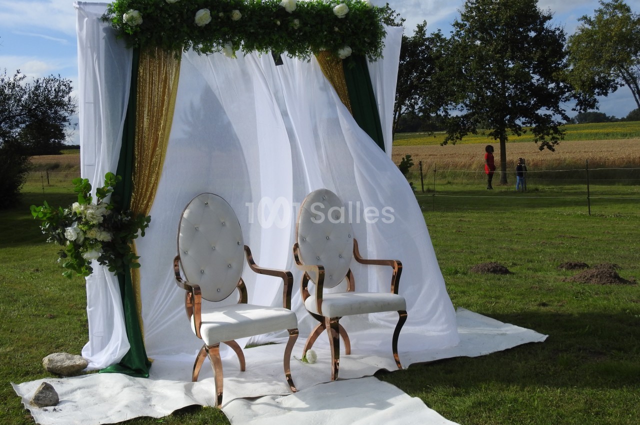 Deux fauteuils blancs devant une arche décorée de voilages, fleurs et feuillage, installée sur une pelouse.