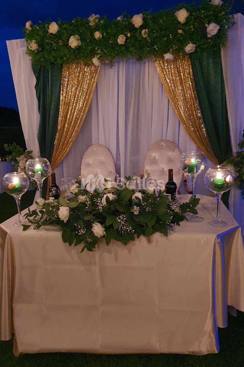 Table décorée pour un mariage avec nappes blanches, chandeliers, fleurs blanches et arche ornée de rideaux dorés et verts.