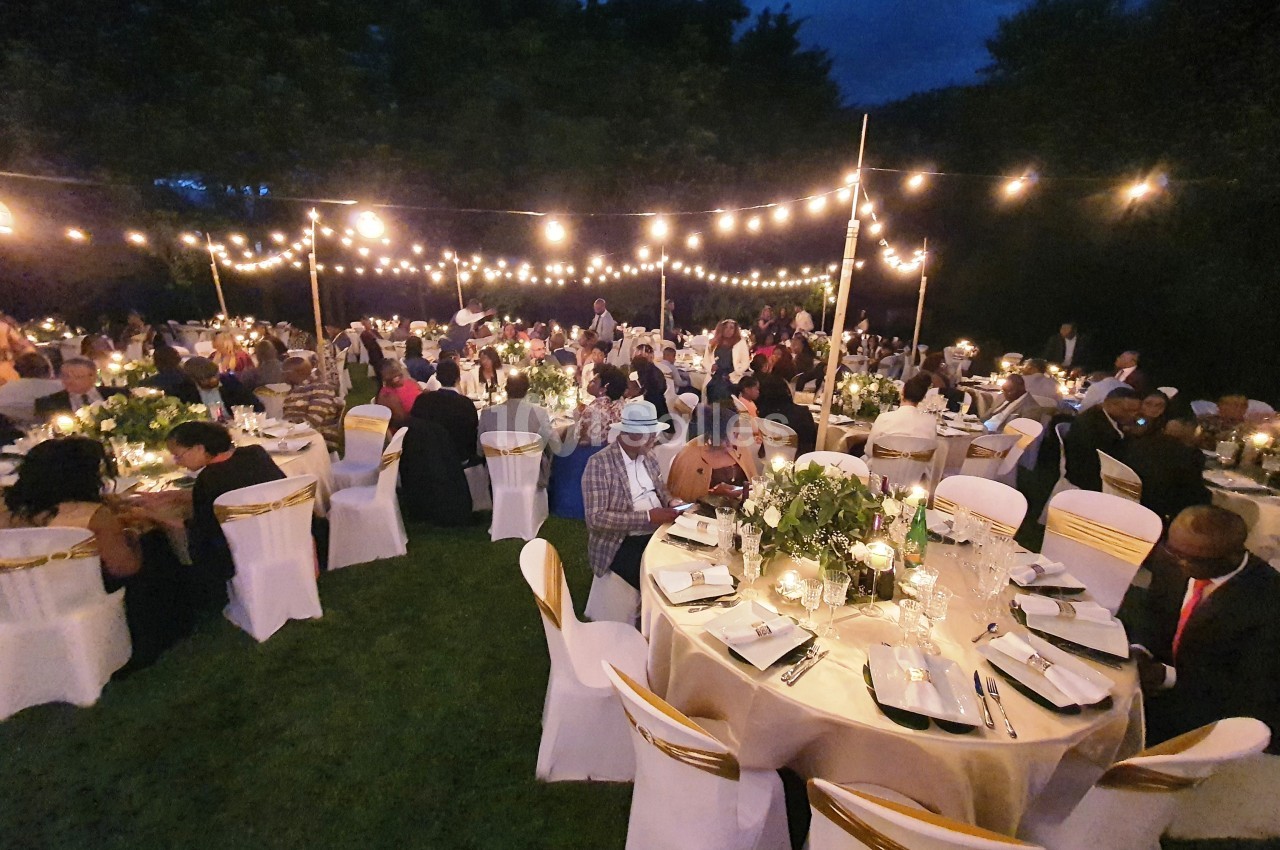 Invités assis à des tables décorées sous des guirlandes lumineuses dans un jardin en soirée.