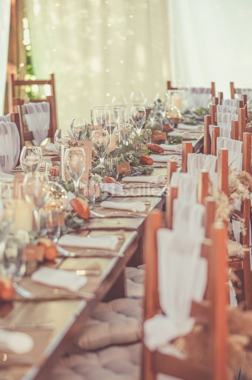 Table de banquet décorée avec des verres, des bougies et des éléments végétaux, entourée de chaises en bois.