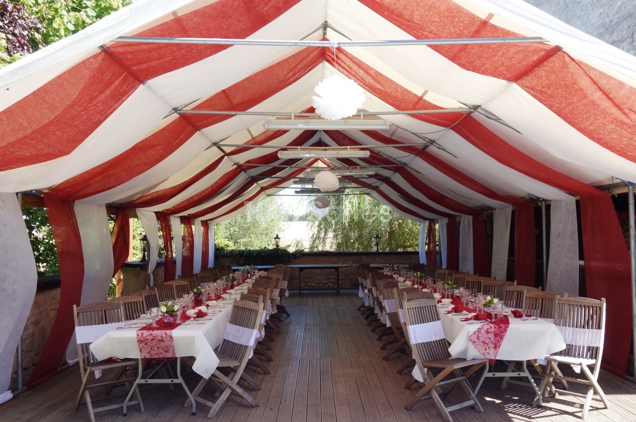 Tente rouge et blanche abritant des tables dressées avec nappes et décorations pour un repas en extérieur.