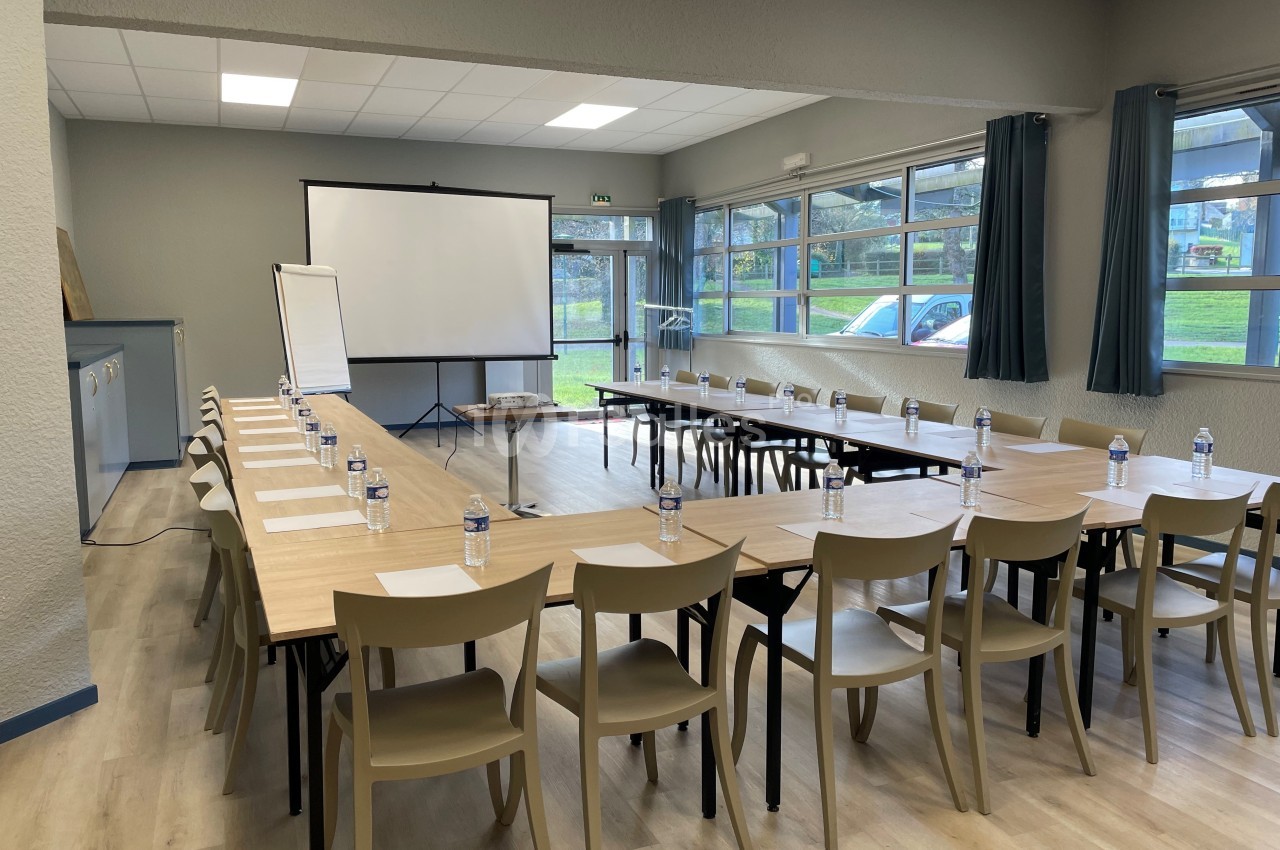 Salle de réunion lumineuse avec tables disposées en U, chaises, tableau blanc et écran de projection.