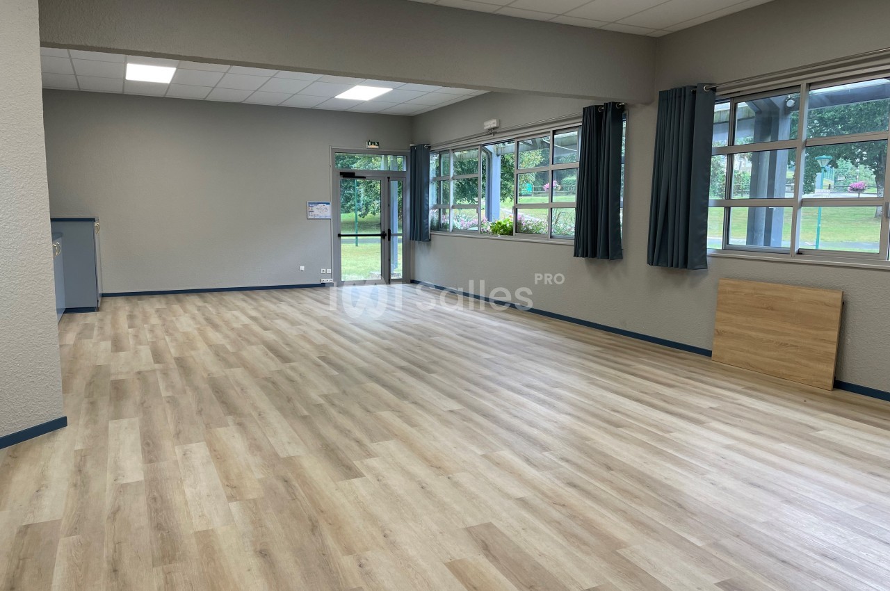 Salle vide avec parquet clair, murs gris, grandes fenêtres laissant entrer la lumière naturelle et vue sur un espace vert.