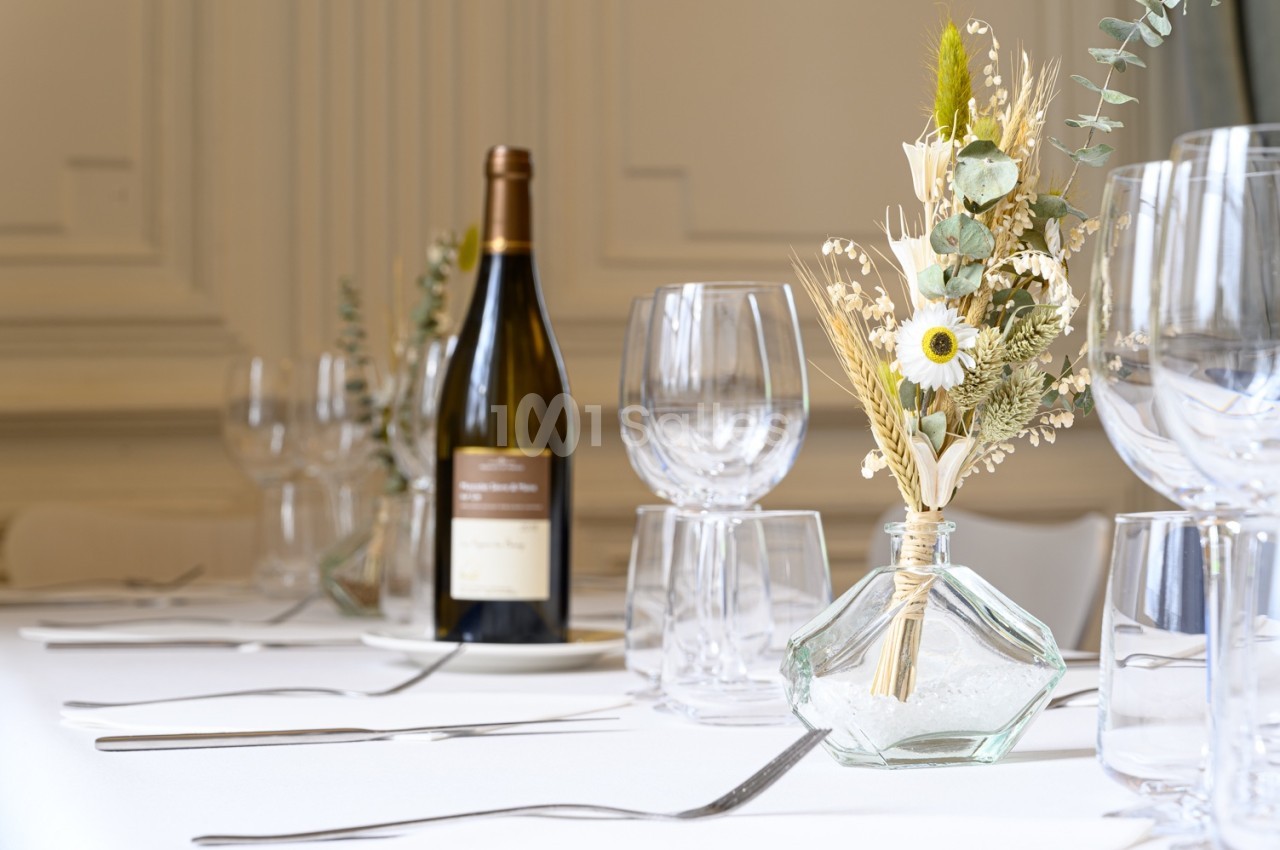 Table dressée avec une bouteille de vin, des verres et un bouquet de fleurs séchées dans un vase en verre.
