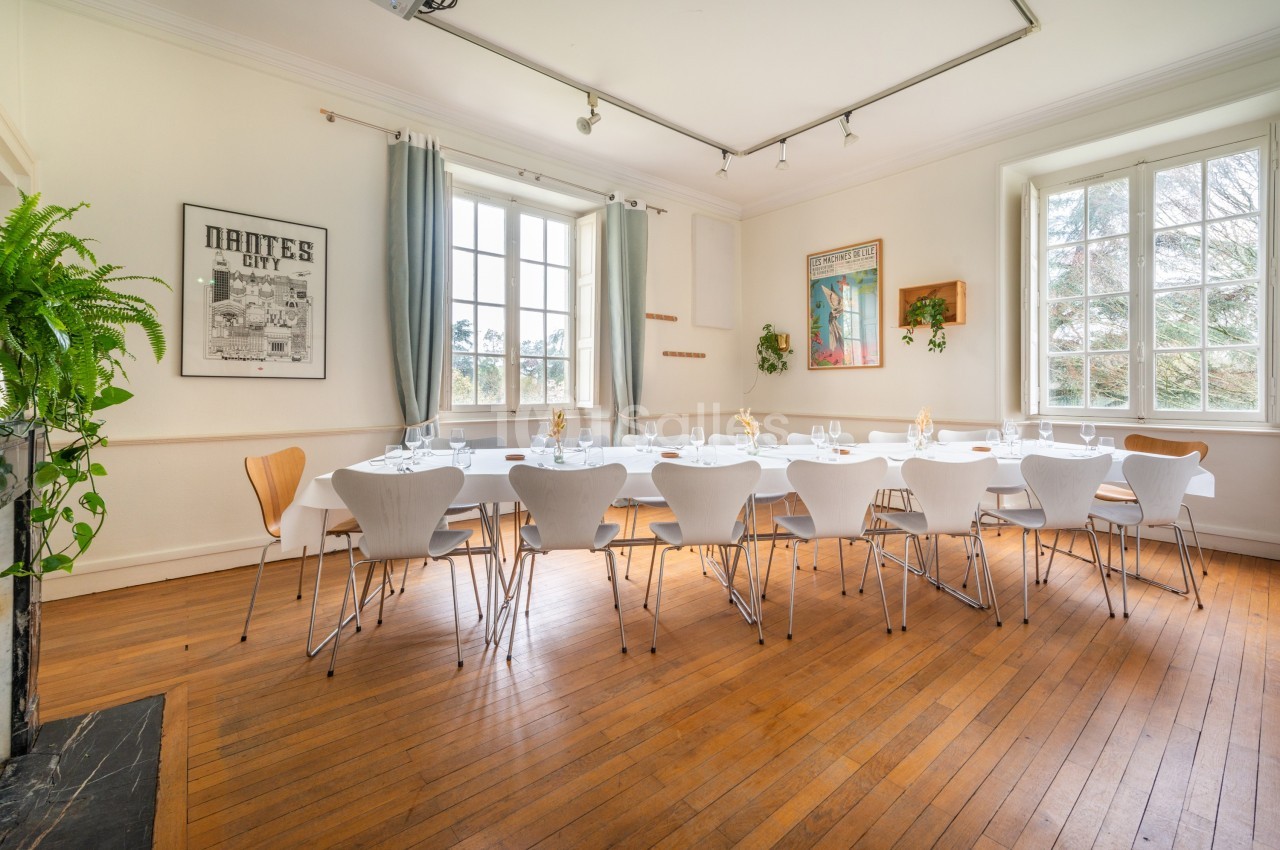 Salle lumineuse avec grande table dressée, chaises modernes, parquet en bois et fenêtres donnant sur un extérieur verdoyant.