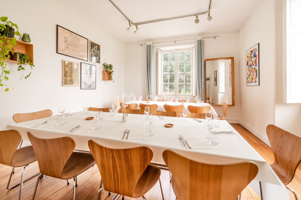 Salle lumineuse avec une grande table dressée, chaises en bois, décorations murales et grande fenêtre donnant sur l…