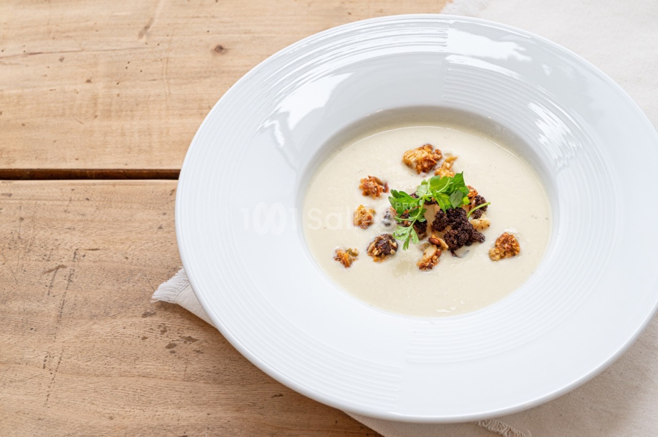 Assiette de velouté crémeux garnie de croûtons dorés, herbes fraîches et miettes croustillantes, sur une table en bois.