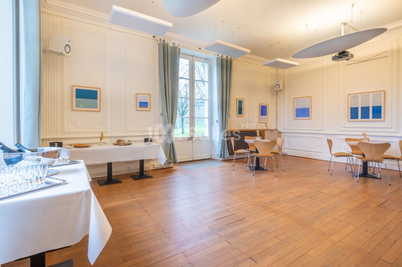 Salle lumineuse avec parquet, tables et chaises en bois, murs ornés de cadres, et buffet dressé près des fenêtres.