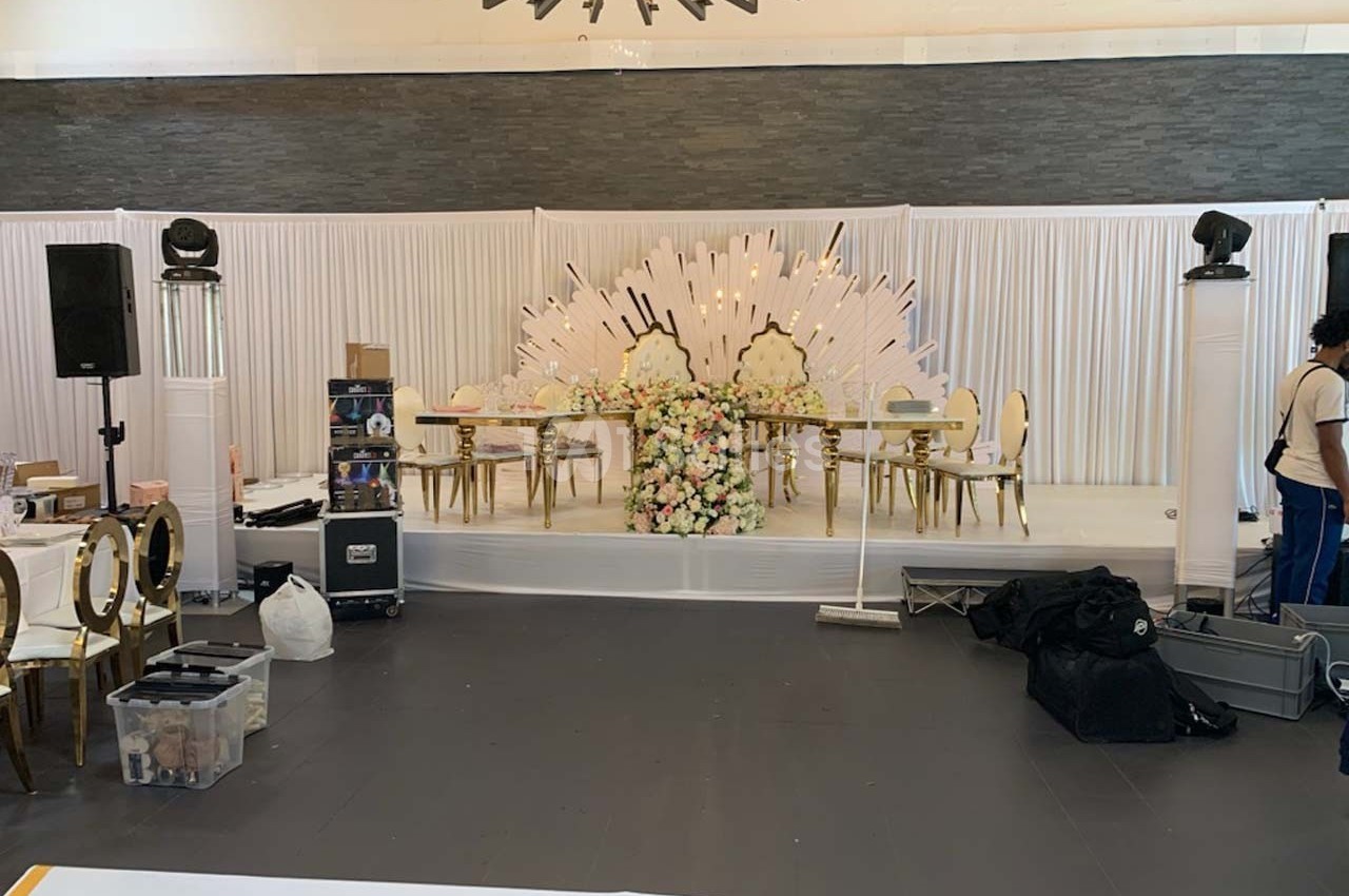 Scène décorée pour un événement avec une table ornée de fleurs, des chaises dorées et un fond blanc lumineux.