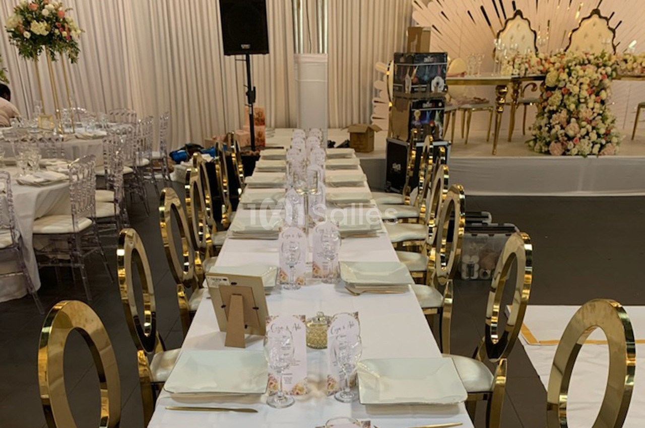 Table de réception élégamment dressée avec chaises dorées, nappes blanches et décor floral en arrière-plan.