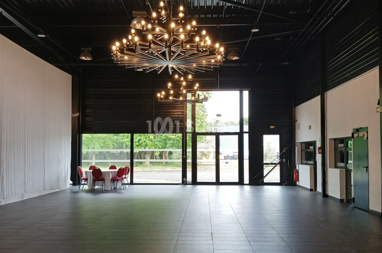 Salle spacieuse avec sol en carrelage, grand lustre central, baies vitrées et quelques chaises rouges près d'une table.