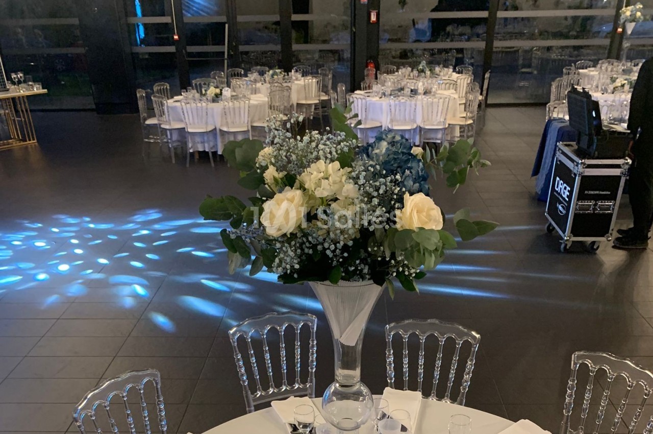 Centre de table floral avec roses blanches et feuillage, dans une salle de réception élégante avec tables dressées.