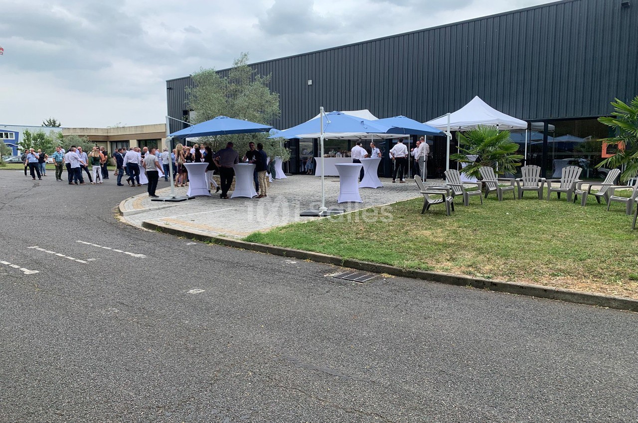 Groupe de personnes rassemblées sous des tentes blanches et bleues dans une cour extérieure près d'un bâtiment industriel.