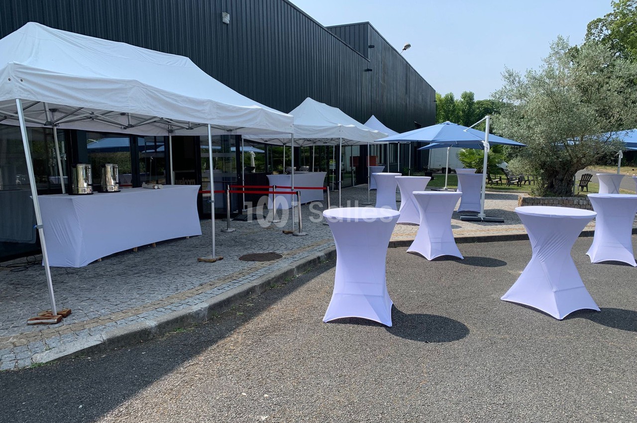 Tentes blanches et tables hautes dressées en extérieur sur un sol pavé devant un bâtiment sombre.