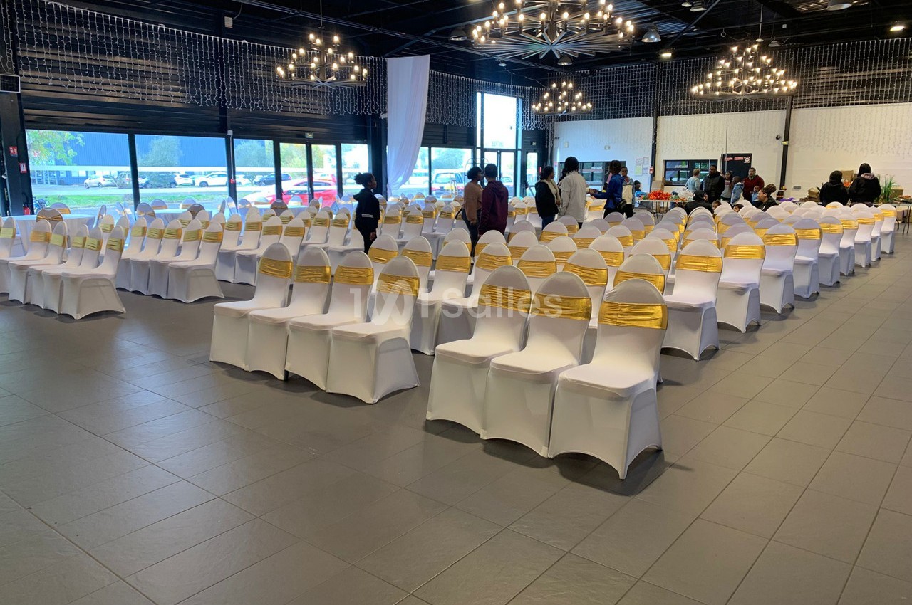 Salle de réception avec des rangées de chaises blanches ornées de rubans dorés, préparée pour un événement.