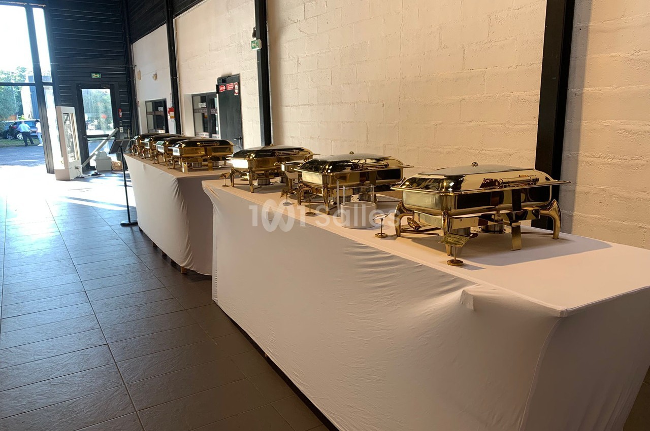 Buffet avec plusieurs plats en inox sur des tables recouvertes de nappes blanches dans une salle lumineuse.