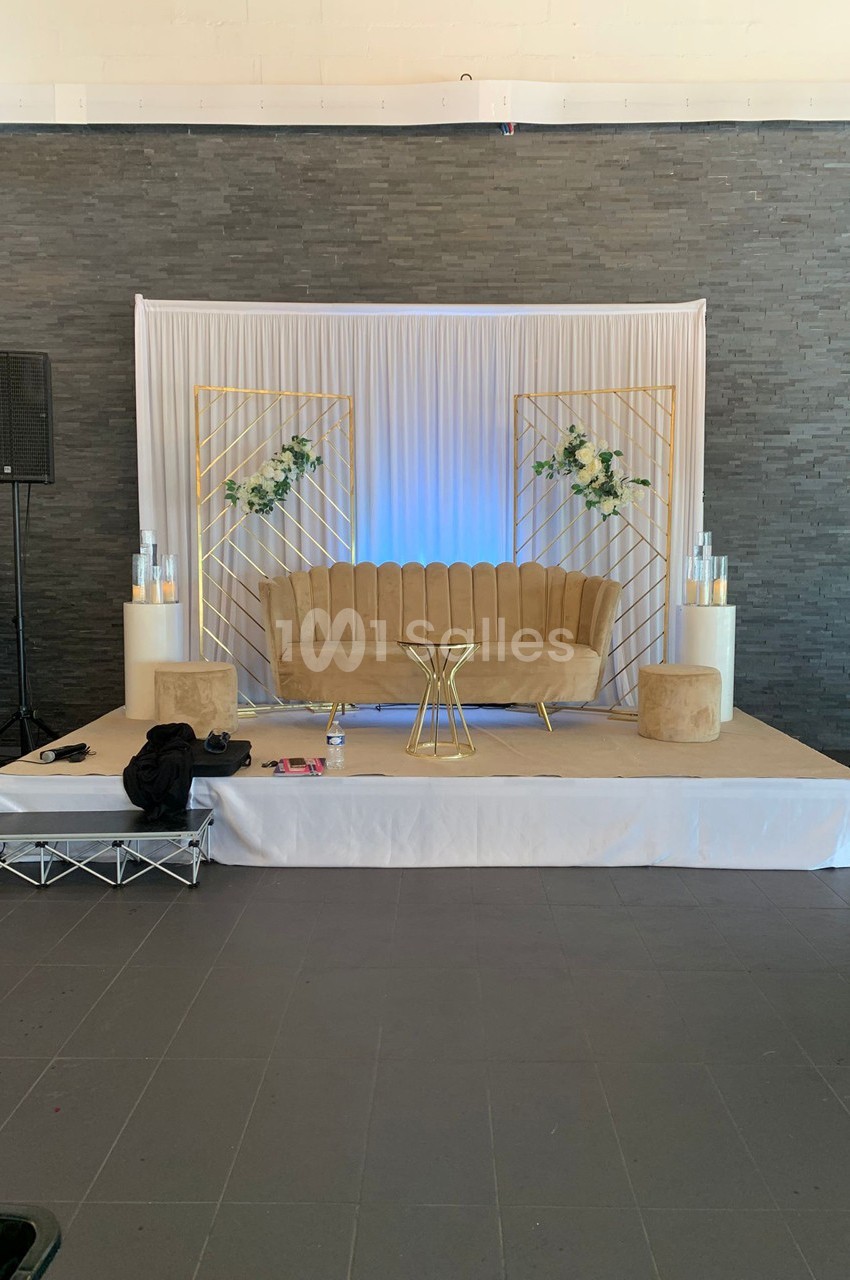 Canapé beige avec table dorée, décor floral et rideaux blancs sur une scène avec fond en pierre grise.