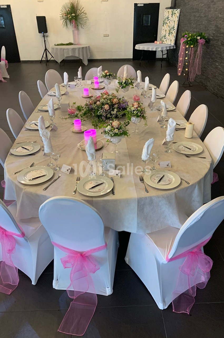 Table ovale décorée pour un événement, avec nappes blanches, chaises ornées de rubans roses et centre de table floral.